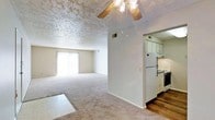 This single-floor space offers a practical and functional layout connecting the main living area to the kitchen. The living room is carpeted and features a textured ceiling with a ceiling fan fitted with a light fixture, providing both air circulation and ambient lighting. A sliding glass door along the far wall fills the room with natural light and hints at outdoor access, while vertical blinds offer privacy when needed. A small tiled section near the center of the living area provides a subtle transition point within the space. The kitchen is partially open to the living area, separated by a short dividing wall that maintains a sense of connection between the two spaces. White cabinetry lines the kitchen walls, complemented by hardwood-style flooring that contrasts with the carpet in the living room. Standard appliances, including a refrigerator and oven range with an overhead ventilation hood, are present. The overall color palette is neutral, with beige tones throughout, creating a clean and understated ambiance. The layout is straightforward and well-suited for everyday living.
