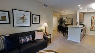 This single-floor residence offers a thoughtfully designed open-concept layout that seamlessly connects the living, dining, and kitchen areas, creating a cohesive and functional living environment. The kitchen is outfitted with warm medium-toned cabinetry, stainless steel appliances including a refrigerator and microwave, and a light-toned countertop peninsula that doubles as a breakfast bar with seating for additional guests. Recessed and pendant lighting illuminate the space with a warm, inviting glow. Adjacent to the kitchen, the dining area accommodates a glass-top table surrounded by dark upholstered chairs, anchored by a stylish pendant light overhead. The living room extends naturally from the dining space, featuring neutral wall tones, plush carpeting, and elevated ceilings that lend an open, airy feel. A sleek leather sofa, framed wall art, and carefully placed accent lighting contribute to the room's refined yet comfortable ambiance. An open doorway leads to a bedroom visible in the background, hinting at a well-proportioned sleeping area. Throughout the space, a cohesive neutral color palette, plush carpeting, and tasteful decor create a warm, welcoming atmosphere suited for both everyday living and entertaining.