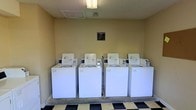 This communal laundry room is a practical, well-organized facility designed to serve the needs of multiple users. The room is equipped with a row of four top-loading washing machines, each coin-operated and positioned side by side along the back wall, providing ample capacity for shared use. The machines are uniformly white and well-maintained, contributing to a clean and functional environment. Overhead, a flush-mounted fluorescent light fixture provides bright, even illumination throughout the space. The flooring features a classic black-and-white checkered tile pattern that adds a touch of visual interest to the utilitarian setting. The walls are painted in a neutral yellow-beige tone, keeping the atmosphere light and open. A posted notice board is mounted on the back wall above the machines, likely displaying laundry room rules and usage guidelines for residents. Ventilation is provided via ceiling vents, ensuring the space remains comfortable during use. Overall, this laundry room is a straightforward, efficiently laid-out shared facility that prioritizes functionality and ease of use for its occupants.
