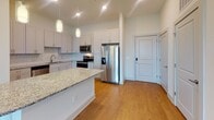 Located at 1715 Legion Rd in Chapel Hill, North Carolina, this well-appointed single-floor residence combines modern finishes with a thoughtfully designed layout. The kitchen is a standout feature, showcasing white shaker cabinetry extending to the ceiling, granite countertops, a decorative tile backsplash, and a full suite of stainless steel appliances including a French door refrigerator, electric range, and over-the-range microwave. Pendant lighting and a central island add both function and style, while warm hardwood-style flooring ties the space together seamlessly into the adjacent living area. The living area is bright and open, with large windows fitted with horizontal blinds that invite natural light and offer views of the surrounding neighborhood. The bedrooms are carpeted for comfort and feature generous windows with pleasant outdoor views of mature trees and the surrounding community. The primary bathroom is elegantly finished with a dual-sink granite vanity, brushed nickel hardware, a large mirror, and a glass-enclosed tile shower. Clean white walls and consistent trim work throughout the unit create a cohesive, contemporary aesthetic that is both inviting and functional.