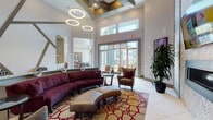 This thoughtfully designed amenity space offers a dynamic blend of social and functional areas across a single level. The centerpiece is a dramatic main lounge boasting soaring double-height ceilings adorned with a coffered detail and striking circular pendant light fixtures. Floor-to-ceiling windows and transom windows flood the space with natural light while offering views of the surrounding exterior. The lounge is furnished with a curved burgundy sectional sofa, leather ottoman, and complementary armchairs arranged atop a bold patterned area rug, creating an inviting, upscale atmosphere.

Adjacent areas transition seamlessly through open corridors featuring warm wood-clad columns, large-format tile flooring, and light hardwood accents. A glass-enclosed conference or co-working room provides a quiet, professional setting. A casual café-style seating area with high-top tables and modern bar stools sits beside floor-to-ceiling windows with sheer drapery, ideal for informal gatherings.

A game room with a billiards table and additional lounge seating adds a recreational dimension. Throughout the space, curated contemporary artwork, textured accent walls, indoor greenery, and a sleek wall-mounted fireplace contribute to a cohesive, vibrant aesthetic.