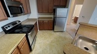 This single-story space includes a well-equipped kitchen that serves as a central feature of the home. The kitchen is outfitted with rich, medium-toned wood cabinetry that extends to the upper walls, providing generous storage space. The cabinetry is complemented by light granite-style countertops that offer ample workspace for meal preparation. Appliances include a black electric coil-burner range with oven, a stainless steel refrigerator, and a built-in microwave, offering a practical and cohesive setup. The flooring throughout the kitchen consists of neutral, stone-patterned ceramic tiles that are easy to maintain and add a clean, classic look to the space. A glimpse into the adjoining area reveals carpeted flooring, suggesting a seamless transition into a living or dining space. The open sightline between the kitchen and the adjacent room contributes to an interconnected, accessible layout. The overall design balances functionality with a warm aesthetic, making it a practical space for everyday living.