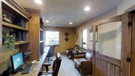 This single-floor space showcases a warm, rustic aesthetic with rich wood tones and earthy wall colors that create an inviting, lodge-inspired atmosphere. The room is thoughtfully divided into two functional zones: a dedicated workspace and a casual seating area. The workspace features a built-in wooden desk running along one wall, complemented by a tall wooden cabinet with crown molding detail, providing ample storage. Task-oriented office chairs complete the workstation setup. The seating area is anchored by supple brown leather armchairs arranged around a wooden coffee table, offering a comfortable space for conversation or relaxation. Natural light filters in through multiple windows on adjacent walls, including one with slatted blinds, brightening the interior while maintaining privacy. Recessed ceiling lighting supplements the natural light throughout. Decorative wall accents, including diamond-shaped ornaments and what appear to be snowshoes, reinforce the rustic theme. Light wood-toned flooring with subtle striping ties the space together, while wood-paneled wall sections add textural depth and cohesion to the overall design.