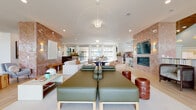 This single-floor space presents a thoughtfully designed open-plan layout that seamlessly integrates multiple functional zones within a bright, contemporary setting. Light wood-look tile flooring runs throughout, unifying the space while contributing to its warm, airy character. Large black-framed windows and glass doors flood the interior with natural light and provide views of the surrounding outdoor landscape.

The expansive lounge area anchors the main living zone, featuring a statement pendant chandelier with a dramatic radial design overhead. Comfortable seating arrangements are complemented by a wall-mounted fireplace set within a textured terracotta tile surround, adding warmth and visual depth. A mounted television sits above the fireplace, creating a media-friendly gathering space.

A dedicated dining area showcases a substantial travertine-toned table surrounded by upholstered chairs with brass-finished legs, set beneath the same striking chandelier. Nearby, a welcoming reception area features fluted sage-green desk panels topped with richly veined stone countertops, illuminated by gold pendant lights.

The kitchenette is outfitted with white cabinetry, brass hardware, a marble-look countertop, a terracotta zellige tile backsplash, and an undercounter beverage cooler. Throughout, the space maintains a cohesive, sophisticated aesthetic with curated décor and abundant natural light.