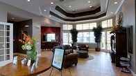 This single-story space functions as a polished lobby or leasing office, designed to make a strong first impression. The most prominent architectural feature is the multi-tiered tray ceiling, accented with dark trim detailing and recessed lighting that adds depth and a refined, upscale character to the room. Large windows along the exterior wall flood the space with natural light while offering views to the outdoors, enhancing the open and airy atmosphere.

The main area is furnished with rich brown leather seating, including armchairs and an ottoman arranged on a area rug, creating an inviting and comfortable lounge space for visitors. Tall potted trees flank the window wall, adding a natural, organic element to the interior design.

Adjacent to the main seating area is a secondary space with a darker accent wall in deep red and a service counter or bar area with seating, suggesting a waiting or hospitality zone. Light-toned tile flooring runs throughout, complementing the neutral gray walls and dark ceiling accents. A reception or display table in the foreground, along with informational signage, reinforces the professional and welcoming nature of this well-appointed space.