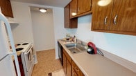 This single-floor unit is thoughtfully laid out to maximize functionality and comfort throughout its living spaces. The kitchen is equipped with warm medium-toned wood cabinetry, both upper and lower, providing ample storage. A neutral laminate countertop runs along the workspace, housing a stainless steel double-basin sink and accommodating appliances such as a range and dishwasher. Light tile flooring and a textured ceiling complete the kitchen's practical aesthetic, and an open passageway connects it to the adjacent living area.

The living and dining space is generously sized, finished with plush wall-to-wall carpeting in a neutral beige tone. A large window allows natural light to fill the room, while a pendant light fixture serves as a simple focal point. The textured ceiling is consistent throughout the unit, contributing to a cohesive look.

The bedroom is bright and open, featuring carpet flooring, a window dressed with horizontal blinds, and a louvered closet door providing accessible storage. Multiple HVAC vents ensure climate control throughout.

The bathroom offers a white vanity with a wide mirror framed by overhead lighting, a tiled backsplash, and clean, bright finishes that keep the space feeling fresh and well-maintained.