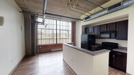 This single-floor unit showcases a compelling industrial aesthetic rooted in its original warehouse or converted factory architecture. The open-concept living and kitchen area is defined by soaring concrete ceilings with exposed wooden planks, visible ductwork, and utility pipes — all hallmarks of authentic loft-style construction. Large, grid-paned factory windows dominate the living area, flooding the space with natural light and offering views of the surrounding urban streetscape. Original brick walls add warmth and texture, providing a striking contrast against the neutral painted surfaces throughout.

The kitchen is positioned along one wall and separated from the living area by a peninsula counter with an integrated sink. Dark espresso cabinetry is paired with black appliances, including a refrigerator, electric range, and built-in microwave, creating a bold, cohesive look. Wood-tone laminate flooring runs throughout the main living spaces, complementing the raw industrial elements overhead.

A separate bedroom features neutral gray walls, dark hardwood-style flooring, and exposed ductwork consistent with the rest of the unit. The bathroom includes a pedestal sink with a vanity mirror, white tile wainscoting, and a full tub-shower combination, offering clean and functional finishes throughout.