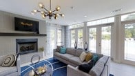 Located at 1910 Capital Creek Dr in Wake Forest, NC, this single-floor space is designed with a modern aesthetic and a welcoming ambiance. The lounge area serves as the centerpiece of the space, featuring a sleek, tile-surround fireplace flanked by a wall-mounted television, creating an inviting focal point. A striking multi-globe chandelier with an industrial-modern design hangs overhead, adding a bold decorative element to the bright, white ceiling. Comfortable gray sofas and sectionals are arranged around a vivid cobalt blue area rug, accented with colorful throw pillows in teal, mustard, and navy tones that add personality and warmth. Large floor-to-ceiling windows and French doors wrap around the room, flooding the interior with generous natural light and offering views of the outdoor surroundings. Sheer patterned curtains frame the windows, softening the light while maintaining an open, airy feel. The overall design balances contemporary style with comfort, making this a functional and visually appealing gathering space. The abundance of windows and thoughtful furnishings create an environment that feels both polished and approachable, well-suited for community engagement or social interaction.