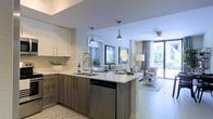 This contemporary single-floor residence showcases a thoughtfully designed open-concept layout that seamlessly blends the kitchen and living areas into one cohesive, functional space. The kitchen is a standout feature, boasting two-toned cabinetry with warm wood lower cabinets and crisp white upper cabinets, complemented by clean white quartz countertops. A peninsula island provides additional workspace and casual dining, accented by elegant metallic pendant lights that hang overhead. Stainless steel appliances, including a dishwasher, add a modern touch throughout. The living area flows naturally from the kitchen, bathed in abundant natural light streaming through large windows and a glass door that opens to an outdoor space, offering views of lush greenery beyond. A ceiling fan overhead adds both style and practicality to the living zone. Neutral tile flooring runs throughout, enhancing the sense of openness and continuity. Tasteful décor, including framed artwork and layered lighting from floor lamps, creates a warm and inviting ambiance. The overall atmosphere is bright, airy, and modern, reflecting a refined yet approachable residential aesthetic.