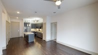 This modern single-floor residence in Dallas offers a thoughtfully designed open-concept layout that seamlessly connects the living and kitchen areas. The kitchen is a standout feature, showcasing dark gray cabinetry, a classic subway tile backsplash, and a full suite of stainless steel appliances, including a French door refrigerator, range, and dishwasher. A granite-topped island provides additional prep space and serves as a natural gathering point between the kitchen and living area. Pendant lighting over the island adds a refined touch, complementing the ceiling fans that ensure comfortable airflow throughout the space. Rich, dark hardwood flooring runs continuously across the entire floor plan, grounding the space with warmth and visual continuity. The clean white walls and neutral palette create a bright, open atmosphere that balances the darker tones of the cabinetry and flooring. Generous ceiling height enhances the overall sense of openness. A hallway off the main living area leads to additional rooms, offering a practical and well-organized flow. The overall design reflects a contemporary aesthetic with quality finishes suited for modern urban living in Dallas.