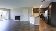 This single-floor space showcases a thoughtfully designed open-concept layout that seamlessly connects the living area and kitchen, creating a sense of spaciousness and ease of flow. The living area is anchored by a clean, white-framed fireplace that adds warmth and a classic focal point to the room. Rich, dark hardwood floors run throughout, providing an elegant contrast to the neutral, light-toned walls and crisp white trim and baseboards.

The kitchen is outfitted with dark espresso-toned cabinetry that offers generous storage, complemented by a light stone countertop that extends into a peninsula with a built-in sink — ideal for casual dining or food preparation. Stainless steel appliances, including a range and an over-the-range microwave, add a modern, polished finish to the culinary space.

Recessed lighting in the kitchen ensures the area is well-lit and functional, while a ceiling fan contributes to comfortable air circulation throughout. The overall ambiance of the space is clean, contemporary, and move-in ready, with a neutral palette that provides a versatile backdrop for a variety of interior design styles.