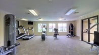 This fitness center offers a clean, functional layout designed to support a well-rounded workout routine. The space is finished with neutral-toned walls and durable carpet tile flooring, creating a practical and comfortable environment for exercise. Overhead fluorescent lighting ensures the room is consistently and evenly lit throughout.

A row of large windows along the far wall allows natural light to fill the space and provides pleasant views of the surrounding outdoor greenery, contributing to an open and inviting atmosphere. A wall-mounted flat-screen television is positioned toward the front of the room, offering entertainment during workouts.

The cardio area is well-appointed, featuring treadmills and an elliptical trainer positioned to take advantage of the natural light and outdoor views. A stationary bike provides an additional low-impact cardio option. For strength training, a multi-station cable machine and an accompanying bench offer versatile resistance workout capabilities.

The generous open floor space between and around the equipment allows for comfortable movement and accommodates a variety of exercise routines. The overall layout is thoughtfully organized, making efficient use of the available space while maintaining a clean and uncluttered feel. This fitness center is well-suited for residents or guests seeking convenient access to essential exercise equipment.