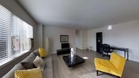 This single-floor space offers a clean, modern aesthetic centered around an open and airy living area. The room is finished with smooth light-toned hardwood flooring that runs throughout, complementing the crisp white walls and flat white ceiling that together create a bright, welcoming atmosphere. Natural light filters in through a window along one wall, adding warmth to the interior.

The living area is thoughtfully arranged with a plush gray sectional sofa accented by vibrant yellow and patterned throw pillows, adding a pop of color to the otherwise neutral palette. A sleek black coffee table with glass candle accents serves as the focal centerpiece, flanked by a yellow upholstered ottoman that reinforces the bold accent theme. A flat-screen television sits atop a low-profile black media console, accompanied by a framed mirror mounted on the wall behind it.

A tall black display cabinet in the corner provides additional storage and decorative display space. The open layout allows for generous circulation and a sense of spaciousness, with a doorway visible leading further into the home. The overall ambiance is contemporary and inviting, balancing functional living with a cohesive, stylish design sensibility.