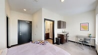 Located at 916 N College Ave in Bloomington, Indiana, this efficiently designed unit offers two bedrooms and one bathroom across a single floor. The primary bedroom is generously proportioned relative to the overall footprint, featuring light-toned tile flooring, crisp white walls, and multiple windows fitted with blinds that allow for ample natural light and outdoor views. A wall-mounted HVAC unit provides individual climate control, while a dedicated study area with a desk and chair makes the room functional for both rest and productivity. Built-in under-bed drawers offer additional storage convenience. A walk-in closet with wire shelving and a built-in dresser unit provides organized storage space. The kitchenette area within the unit includes dark espresso cabinetry, granite-style countertops, and a compact refrigerator, offering essential in-room conveniences. The private bathroom is well-appointed with matching dark cabinetry, a granite-topped vanity with an undermount sink, a large mirror with overhead lighting, and a walk-in shower. Dark-framed doors throughout the unit add a modern, contemporary contrast to the neutral color palette, creating a cohesive and polished aesthetic throughout.