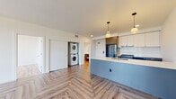 Located at 4055 Mercantile Dr in Lake Oswego, Oregon, this thoughtfully designed single-floor residence combines modern finishes with practical living. The open-concept layout is anchored by striking herringbone-patterned hardwood flooring that flows seamlessly throughout the space, creating a sense of continuity and warmth.

The kitchen is a standout feature, outfitted with two-tone cabinetry in white and taupe-gray, a bold blue kitchen island with a white quartz countertop, brass-accented fixtures, a deep undermount sink, stainless steel appliances including a gas range and French door refrigerator, and a classic subway tile backsplash. Pendant lighting adds a refined touch overhead.

The living area is bright and airy, benefiting from a large bank of floor-to-ceiling windows that flood the space with natural light and offer views of the surrounding Lake Oswego neighborhood. A wall-mounted mini-split system provides climate control throughout.

Two bathrooms serve the residence: one with a tiled tub-shower combination and the other with a spacious glass-enclosed walk-in shower, both featuring gray vanity cabinetry and light wood-tone flooring. The unit also includes a dedicated in-unit stacked washer and dryer closet for added convenience. Bedrooms are generously sized with ample natural light and closet space.