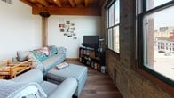 Located at 114 W Oregon St in Milwaukee's vibrant Walker's Point neighborhood, this single-floor loft showcases a compelling blend of industrial heritage and comfortable modern living. The space is defined by its striking architectural character, including exposed heavy timber ceiling beams, original brick walls, and warm hardwood flooring that flow throughout the open layout.

The living area is generously proportioned, bathed in natural light from large windows that frame views of the surrounding cityscape. The exposed beam ceiling adds dramatic height and a sense of openness, while the raw brick walls provide rich texture and a distinctly urban aesthetic.

The kitchen features warm wood cabinetry, light stone-toned countertops, and a double stainless steel sink. Exposed brick serves as the backsplash, tying the kitchen seamlessly into the loft's industrial character. Stainless steel appliances, including a range, microwave, and dishwasher, complete the functional space.

The bedroom continues the loft's signature aesthetic with exposed timber beams framing the room, complemented by carpeted flooring for added comfort. A window provides natural light to the restful retreat.

The bathroom is clean and well-appointed, featuring a vanity with a large mirror and a shower/tub combination. Throughout the space, the interplay of raw industrial materials and warm finishes creates an inviting, distinctive atmosphere ideal for urban living in Milwaukee.