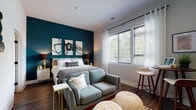 This thoughtfully designed studio offers a seamless open-concept layout that efficiently combines living, sleeping, and dining functions into one cohesive space. The sleeping area serves as a focal point, featuring a bold teal accent wall adorned with abstract artwork, large windows dressed with sheer white curtains and blinds that invite generous natural light and views of surrounding greenery. A cozy loveseat, woven poufs, and a bistro-style bar table complete the multifunctional living zone.

The kitchen is well-appointed with two-toned cabinetry — white uppers paired with dark wood-grain lowers — complemented by a neutral tile backsplash, white quartz-style countertops, a black matte sink fixture, and a full suite of stainless steel appliances including a refrigerator, electric range, and over-the-range microwave. Track lighting overhead adds a contemporary touch to the open living area.

The bathroom maintains a clean, modern aesthetic with a floating wood-toned vanity, a wide mirror, matte black hardware accents, and botanical-themed wall art. A tub-shower combination and warm wood-look flooring — consistent throughout the entire space — round out the refined, contemporary feel of this efficiently designed studio.