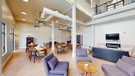 The Stoney Creek Community Room at 2710 Wilkie Dr in Rapid City, South Dakota, is a generously sized, multi-functional gathering space designed to serve residents with both comfort and practicality. The room's most striking architectural feature is its soaring double-height ceiling, which creates an airy, open atmosphere throughout. Exposed ductwork and structural columns add an industrial character, while light hardwood flooring and a neutral color palette keep the space feeling warm and inviting.

The main area is thoughtfully divided into distinct functional zones. A lounge area is furnished with plush, mid-century modern sofas and accent chairs arranged around round wooden coffee tables, ideal for casual conversation or relaxation. Nearby, a wall-mounted television flanked by two armchairs provides a dedicated media corner. The dining area accommodates multiple wooden tables and chairs, with large windows offering direct views of the outdoor pool and surrounding greenery, filling the space with abundant natural light.

A fully equipped kitchen features rich wood cabinetry, dark granite countertops, a mosaic tile backsplash, stainless steel appliances, and a breakfast bar with seating. Ceiling fans are distributed throughout to ensure comfortable airflow. A staircase with sleek black metal railings leads to an upper-level overlook balcony, adding vertical dimension and architectural interest to this versatile community space.