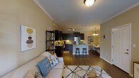 This single-floor space is thoughtfully designed with an open-concept layout that integrates the living room, kitchen, and dining area into one cohesive, flowing environment. Crown molding runs along the ceiling throughout, adding a refined architectural detail that elevates the overall aesthetic.

The living area is anchored by a plush sofa and a textured area rug, creating a comfortable and welcoming atmosphere. Warm yellow-toned walls contribute to an inviting ambiance across the space.

The kitchen is a standout feature, equipped with rich, dark espresso cabinetry that contrasts elegantly with granite countertops and stainless steel appliances. A peninsula with bar-stool seating serves as both a functional workspace and a social hub, bridging the kitchen and living areas. Track lighting illuminates the kitchen, ensuring ample task lighting.

Adjacent to the kitchen, the dining area benefits from the warm glow of a chandelier overhead, making it an ideal setting for meals and gatherings. The continuous hardwood-style flooring ties each zone together, reinforcing the sense of openness and cohesion throughout the space.