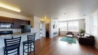 This single-floor unit offers a thoughtfully designed open-concept layout that connects the kitchen, living, and dining areas into one cohesive and functional space. Rich wood-style flooring runs throughout the main areas, adding warmth and continuity to the design.

The kitchen is equipped with dark espresso cabinetry, granite-style countertops, and a full suite of black appliances, including a refrigerator, stove, dishwasher, and microwave. A breakfast bar with bar seating provides a convenient spot for casual dining and connects the kitchen seamlessly to the living area.

The living room is bright and inviting, featuring a wall of large windows dressed with white blinds that allow generous natural light while offering views of the surrounding neighborhood. The space comfortably accommodates a seating arrangement centered around a coffee table.

A dedicated dining area sits adjacent to the living room, also benefiting from natural light through its own window, and provides ample space for a full dining table and chairs.

The bedroom is a comfortable and well-lit retreat, featuring a large window, carpeted flooring, and enough space for a bed and a dedicated desk area. The full bathroom includes a single-sink vanity with granite-style countertops, a toilet, and a walk-in shower with a glass sliding door, finished with consistent wood-style flooring.