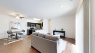 This modern single-floor residence offers a thoughtfully designed open-concept layout that connects the living, dining, and kitchen areas into one cohesive and functional space. The living area is anchored by a plush sectional sofa and features warm hardwood-style flooring that extends throughout, lending a sense of continuity and warmth to the interior. A patterned area rug and a sleek console table with minimal décor add character without overwhelming the clean aesthetic.

The kitchen is well-appointed with dark cabinetry that provides a striking contrast against the white walls and light countertops. Stainless steel appliances — including a refrigerator, microwave, and range — bring a contemporary edge, while the peninsula-style counter creates a natural boundary between the kitchen and the adjoining dining area. The dining space accommodates a small table and chairs, making it ideal for everyday meals.

Recessed lighting and a ceiling fan keep the space bright and comfortable, while the crisp white ceilings enhance the open, airy feel of the interior. The overall ambiance is clean, modern, and welcoming — a well-balanced space that combines practical design with a contemporary residential aesthetic.