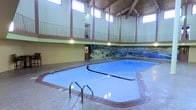This indoor aquatic facility makes a striking impression with its soaring vaulted wood-paneled ceiling and exposed beam construction, creating a warm and inviting atmosphere. Abundant natural light pours in through a row of clerestory windows positioned high along the upper walls, illuminating the space and reflecting off the clear blue water below. The generously sized swimming pool is framed by a tiled surround and features metal entry handrails for easy access, with depth markers visible along the edges. A dramatic, large-scale nature-themed mural depicting a vibrant wildlife and waterscape scene spans the far wall, serving as a bold and memorable focal point. An elevated mezzanine-level walkway with protective railings overlooks the pool area, adding architectural depth and vertical interest to the room. The flooring throughout the surrounding deck area features a durable, textured finish well-suited for a wet environment. Warm ambient lighting complements the natural light, ensuring the space remains bright and welcoming. The overall design balances functionality with aesthetic appeal, making this facility a standout recreational amenity.