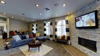 This single-floor space showcases a thoughtfully curated interior that blends bold design choices with comfortable functionality. The main lounge area is anchored by a large navy blue sectional sofa accented with red and yellow throw pillows, arranged around a classic wooden coffee table atop a patterned area rug in yellow and grey tones. Striking black-and-white striped curtains frame a series of tall arched windows that allow generous natural light to fill the room, creating a bright and inviting atmosphere. The combination of hardwood flooring and carpeted zones adds visual variety and warmth underfoot. A prominent stacked stone accent wall serves as a dramatic architectural feature, complemented by recessed ceiling lighting that provides warm, even illumination throughout. Additional seating near the windows includes wicker chairs with colorful cushions, offering a cozy nook for relaxing. In the background, a dining or gathering area is visible, further extending the functional layout of the space. The overall design palette — warm neutrals punctuated by bold yellows, reds, and deep blues — gives the space a vibrant yet grounded character, making it well-suited as a shared lounge or social gathering area.