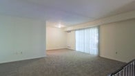 This single-floor residence offers a clean, functional layout centered around a generously sized open living area. The main living space is finished with wall-to-wall carpeting in a neutral grey tone, providing a comfortable and cohesive feel underfoot. Cream-colored walls throughout contribute to a bright, airy ambiance that makes the space feel open and inviting.

A standout feature of the living area is the large sliding glass door dressed with full-height vertical blinds, which allows for abundant natural light to fill the room while also offering direct access to the outdoors. A recessed nook adjacent to the main living area provides a natural opportunity for a defined dining or secondary sitting space, adding versatility to the floor plan.

Practical details include a ceiling-mounted light fixture for general illumination and baseboard heating units positioned along the walls, ensuring year-round comfort. The overall design is straightforward and move-in ready, with a neutral palette that serves as an ideal backdrop for a variety of furnishing styles. The single-floor layout offers ease of accessibility and a seamless flow throughout the home, making it a practical and comfortable living environment.