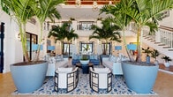 Located at 10535 Hamilton Dawn St in Orlando, FL, this grand lobby space delivers an impressive resort-style atmosphere across a generous single floor. Soaring double-height ceilings define the architectural character of the space, creating an airy, open ambiance that immediately commands attention upon entry. Towering indoor palm trees planted in large decorative containers rise dramatically toward the ceiling, reinforcing the tropical Florida aesthetic throughout.

The main lounge area features a beautifully curated coastal-inspired seating arrangement, with plush sofas and accent chairs in soft blues and whites arranged atop an ornate blue-and-white patterned area rug. Crystal and lantern-style pendant lighting adds a refined touch overhead, complementing the warm-toned herringbone hardwood flooring that flows seamlessly throughout the space.

Black-framed glass doors and tall windows allow abundant natural light to pour in, connecting the interior to the lush outdoor surroundings. A sweeping staircase with rich wood treads, white balusters, and a curated gallery wall of framed artwork serves as a striking architectural focal point. An upper mezzanine level with decorative white railings overlooks the main floor below, adding vertical depth and grandeur. The overall ambiance is one of sophisticated, tropical elegance.