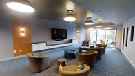 This single-floor communal lounge is designed with a contemporary aesthetic, combining functionality and comfort in a well-appointed gathering space. The room features a neutral color palette of grays and whites, accented by warm earth tones introduced through the varied upholstered seating. A large wall-mounted flat-screen television serves as a focal point, flanked by sleek, low-profile white cabinetry that runs along the wall, providing both storage and a clean architectural line.

The seating arrangement is thoughtfully curated, featuring an eclectic mix of curved barrel chairs and rounded ottomans in shades of brown, tan, and mustard yellow, some accented with patterned throw pillows. This variety encourages both individual relaxation and group interaction. Small accent tables are interspersed throughout, offering practical surfaces within easy reach.

Overhead, a series of modern pendant and flush-mount light fixtures provide warm, evenly distributed illumination throughout the space. A patterned carpet in muted tones of blue and gray anchors the room. Natural light filters in through a window visible at the far end, adding brightness to the overall inviting ambiance.