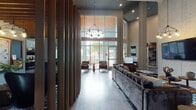This expansive amenity space is defined by its contemporary design and thoughtfully curated zones for socializing, relaxing, and working. The main lounge greets visitors with soaring double-height ceilings, floor-to-ceiling windows that flood the interior with natural light, and a striking chandelier suspended above a seating arrangement of plush velvet sofas and leather accent chairs. Vertical wood slat partitions add architectural warmth and help delineate functional areas throughout the open floor plan.

A prominent concrete panel feature wall serves as a dramatic backdrop in the central lounge, adorned with large-scale abstract artwork and a wall-mounted television. Polished concrete flooring flows consistently throughout, reinforcing the modern industrial aesthetic.

The clubroom kitchen is a standout feature, showcasing a textured stone tile accent wall, multiple wall-mounted flat-screen televisions, rich wood cabinetry, a beverage station, wine refrigerator, and a generous white countertop island with bar seating—ideal for gatherings or casual dining.

A dedicated game and entertainment area features a foosball table, additional seating, and a wall-mounted television beneath a striking slatted wood ceiling detail. Glass doors lead directly to an outdoor patio furnished with seating and ceiling fans, offering a seamless transition between indoor and outdoor amenity spaces.
