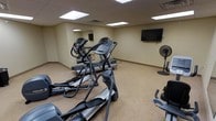 This single-floor fitness room offers a practical and functional workout space designed to accommodate a variety of cardio exercises. The room features a neutral color palette with beige walls and matching low-pile carpeted flooring, creating a clean and utilitarian environment. Overhead, a drop-tile ceiling houses recessed fluorescent panel lighting that evenly illuminates the space.

The room is equipped with multiple cardio machines, including two elliptical trainers and at least one treadmill, providing users with options for varied low- and high-impact workouts. A stationary bike is partially visible, adding further versatility. A large wall mirror on one side of the room enhances the sense of space and allows users to monitor their form during exercise. A wall-mounted flat-screen television is positioned at a comfortable viewing height, offering entertainment during workouts. A standing floor fan provides additional air circulation throughout the room. The overall ambiance is straightforward and purpose-driven, well-suited for regular fitness use.