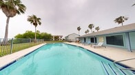 This outdoor pool area offers a well-appointed recreational space designed for relaxation and leisure. The centerpiece is a large, rectangular swimming pool with clear turquoise water, framed by blue tile accents and equipped with sturdy metal entry railings for easy access. The pool's generous size accommodates both casual swimmers and those looking to unwind.

Surrounding the pool is a spacious concrete deck that provides ample room for lounging. The deck is furnished with multiple sun loungers and a seating arrangement featuring a table and chairs, making it ideal for socializing or simply soaking up the outdoors. Tall, mature palm trees are scattered throughout the space, offering a tropical aesthetic and partial shade on sunny days.

The area is enclosed by a metal safety fence, ensuring a secure environment. Lush green grass and additional mature trees line the perimeter beyond the fence, adding to the natural, relaxed feel of the space. A low-profile building with a covered overhang is situated along one edge of the pool deck, providing shelter and additional amenities nearby. The surrounding residential buildings are visible in the backdrop, integrating the pool area seamlessly into the broader community setting.