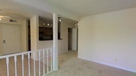 This single-floor space is designed with an open, functional layout that connects its key living areas seamlessly. The main living area features neutral-toned carpeting that extends throughout, creating a cohesive and understated aesthetic. White-painted walls and standard ceiling heights contribute to a clean, simple ambiance.

A white-railed staircase is visible at the entry point, leading up to the main level and adding a classic architectural detail to the space. The kitchen is positioned adjacent to the living area and is partially separated by a half-wall with a raised breakfast bar, providing both division and openness. The kitchen features dark countertops that contrast with lighter cabinetry and is equipped with track lighting for focused illumination.

A doorway visible from the main living area leads to what appears to be a closet or storage space, contributing to the unit's practical storage options. Track lighting fixtures are installed in multiple areas, providing versatile and directed lighting throughout. The tile flooring near the staircase transitions to carpet in the main living space, subtly delineating functional zones within the open floor plan.