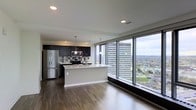 This two-floor residence offers a sophisticated urban living experience defined by clean lines and contemporary finishes throughout. The main living area showcases an open-concept layout where the kitchen flows seamlessly into the living space, creating a generous, light-filled environment. Dark hardwood flooring runs throughout, providing a striking contrast against the bright, neutral walls and white ceiling with recessed lighting.

The kitchen is well-appointed with dark cabinetry, a tiled backsplash, stainless steel appliances including a refrigerator, microwave, and range, and a central island with a light countertop that serves as both a workspace and casual dining area. Pendant lighting hangs above the island, adding a refined touch.

The standout feature of this space is the expansive floor-to-ceiling window wall that floods the interior with natural light and frames dramatic panoramic views of the surrounding cityscape, including high-rise buildings and green spaces below. The elevated position of the unit amplifies these views, creating a strong sense of connection to the urban environment outside.