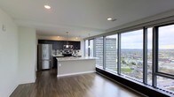 This two-floor residence offers a sophisticated urban living experience defined by clean lines and contemporary finishes throughout. The main living area showcases an open-concept layout where the kitchen flows seamlessly into the living space, creating a generous, light-filled environment. Dark hardwood flooring runs throughout, providing a striking contrast against the bright, neutral walls and white ceiling with recessed lighting.

The kitchen is well-appointed with dark cabinetry, a tiled backsplash, stainless steel appliances including a refrigerator, microwave, and range, and a central island with a light countertop that serves as both a workspace and casual dining area. Pendant lighting hangs above the island, adding a refined touch.

The standout feature of this space is the expansive floor-to-ceiling window wall that floods the interior with natural light and frames dramatic panoramic views of the surrounding cityscape, including high-rise buildings and green spaces below. The elevated position of the unit amplifies these views, creating a strong sense of connection to the urban environment outside.