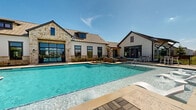 Located in Cypress, Texas, this single-story community amenity center at Bluegrass Park offers a resort-inspired outdoor experience designed for relaxation and social gathering. The centerpiece is a generously sized swimming pool with crystal-clear turquoise water, complemented by an attached spa featuring water jets — all framed by a spacious, light-colored concrete pool deck ideal for lounging.

The clubhouse building presents an attractive blend of natural limestone and white board-and-batten siding, topped with a standing-seam metal roof with dormers. Large glass doors and windows face the pool, allowing abundant natural light to flow through the interior while maintaining a seamless visual connection between indoor and outdoor spaces. A covered patio with bar-height seating extends from one wing of the building, offering an additional gathering spot.

The surrounding outdoor area is thoughtfully appointed with multiple distinct zones: shaded dining tables with large teal market umbrellas provide a casual al fresco setting, while a lush turf lawn area features comfortable wicker lounge sofas with teal accent pillows and a concrete coffee table — perfect for unwinding. Chaise lounges are also positioned poolside for sun-soaking. Black iron perimeter fencing encloses the space, with views of the surrounding neighborhood and open sky beyond.