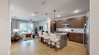 This well-appointed single-floor residence offers a thoughtfully designed open-concept layout that seamlessly connects the kitchen, dining, and living areas. The kitchen is a standout feature, showcasing rich wood-grain cabinetry extending to the ceiling, light quartz countertops, a white subway tile backsplash, black hardware accents, and stainless steel appliances. A central island with a waterfall-edge countertop provides additional workspace and casual seating for multiple guests, illuminated by stylish globe pendant lights overhead. Light wood-tone flooring flows throughout the main living area, enhancing the bright and airy atmosphere. The living room is anchored by large sliding glass doors that open to a private balcony, filling the space with natural light and offering views of the surrounding greenery. A ceiling fan adds both function and style to the living area. The primary bedroom is generously sized with plush carpeting, large windows, and a ceiling fan, creating a comfortable retreat. It connects to a well-appointed en-suite bathroom featuring dual sinks with wood cabinetry, backlit mirrors, large-format tile flooring, a soaking tub, and a glass-enclosed walk-in shower. A dedicated walk-in closet with dual hanging rods and overhead shelving provides ample storage. A secondary living or sitting area with sliding glass doors adds versatility to the floor plan.