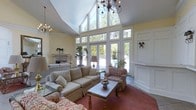 This single-story space makes a striking impression through its living room, which is defined by dramatically vaulted ceilings that rise to impressive heights. A sweeping curved ceiling line adds architectural interest overhead, while an elegant wrought-iron chandelier serves as a focal point above the seating area. A second chandelier echoes the design, reinforcing the refined aesthetic throughout.

The living room is anchored by a wall of floor-to-ceiling windows and glass doors that span the full height of the space, allowing an abundance of natural light to pour in while offering unobstructed views of the surrounding mature trees and outdoor areas. The yellow-toned walls contribute warmth to the bright, airy atmosphere.

A classically styled fireplace with a stone surround adds a traditional focal point to one wall, complemented by decorative wall sconces that enhance the room's layered lighting. The space accommodates a generous arrangement of neutral-toned sofas, patterned accent chairs, and a wooden coffee table, creating a comfortable and inviting seating area. Slate-tiled flooring transitions to rich, deep-red area rugs, grounding the room with warmth and texture.