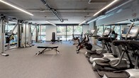 This fitness center is a comprehensively outfitted facility designed to support a wide range of training disciplines. The space features an open-plan layout with a neutral, low-pile carpet flooring throughout, providing a clean and functional foundation for both cardio and strength activities. An exposed ceiling with industrial-style ductwork and linear LED lighting gives the facility a modern, utilitarian aesthetic, while large floor-to-ceiling windows along one wall flood the interior with natural light and offer views of the surrounding greenery outdoors.

The cardio zone is well-stocked with elliptical trainers, stationary bikes, spin bikes, and treadmills, all arranged to maximize space and usability. Mirrored walls extend the visual depth of the room and support form-checking during workouts. A dedicated strength training area features a variety of selectorized machines, including cable systems by Hoist, as well as adjustable benches and a dumbbell rack with a full range of free weights. Functional training equipment including TRX suspension trainers and medicine balls are also present. A separate area with rubberized flooring accommodates heavier strength training with power racks and additional machines. Built-in shelving and storage further support the organization of accessories and equipment throughout the space.