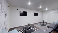 This single-floor space offers a contemporary communal lounge designed with a clean, minimalist aesthetic. The room is finished with crisp white walls and dark hardwood-style flooring, creating a sharp contrast that anchors the modern design. Two flat-screen TVs are mounted side by side along the main wall, making the space well-suited for group viewing or casual entertainment. Recessed and flush-mount ceiling lighting provides consistent, even illumination throughout. The furnishings follow a cohesive black-and-white palette, with bold striped area rugs layered over the dark flooring and low-profile black coffee tables arranged to define separate seating zones. Small potted plants add subtle organic accents against the otherwise neutral backdrop. A security camera is visible near the ceiling, indicating this is a shared or managed facility. A white door on the far wall provides access to an adjacent area. The overall atmosphere is functional yet stylish, reflecting a well-maintained space designed to accommodate multiple occupants comfortably.