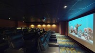 Located at 111 Pearl St in Hartford, Connecticut, this dedicated screening room delivers an immersive, cinema-quality experience within a single, thoughtfully designed space. The room is furnished with rows of plush black leather recliner chairs, each equipped with built-in cup holders, arranged in a tiered layout to ensure unobstructed sightlines to the large projection screen positioned at the front of the room. A ceiling-mounted projector provides crisp, high-quality imagery on the expansive screen, making the space well-suited for film screenings, presentations, or private events. The dark drop-panel ceiling and deep, warm-toned walls in rich burgundy and amber hues create an intimate, distraction-free atmosphere reminiscent of a premium commercial cinema. Strategically placed wall sconces and recessed ceiling lights provide subtle, dimmable ambient illumination that complements the theatrical setting without detracting from the screen. The floor is covered in a bold, multi-colored patterned carpet that adds a dynamic visual element to the otherwise moody interior. Aisle lighting along the stepped floor levels enhances both safety and aesthetics. A dedicated entry door is accessible from the rear of the room, ensuring smooth ingress and egress. Overall, the space offers a polished, fully equipped environment for an elevated viewing experience.