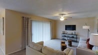 This single-story space centers around a comfortable living room designed for everyday functionality. The room features soft wall-to-wall carpeting underfoot and warm, neutral yellow-toned walls that contribute to an inviting atmosphere. A textured ceiling adds subtle visual depth to the space.

Natural light enters generously through a large sliding glass door dressed with vertical blinds and flanked by decorative curtain panels, creating a bright focal point along one wall. A ceiling fan with an integrated light fixture is centrally positioned overhead, providing both air circulation and ambient lighting.

The living area is arranged around a plush, light-colored sofa facing a wall-mounted flat-screen television supported by an open, industrial-style media console. Rustic wooden stump side tables serve as unique accent pieces, adding a natural, earthy contrast to the otherwise neutral interior. The overall ambiance of the space is casual and lived-in, balancing practical furnishings with simple, approachable design elements suited for comfortable day-to-day living.