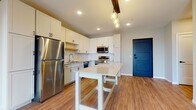 This single-floor residence showcases a thoughtfully designed open-concept layout with warm wood-look flooring flowing throughout the main living areas. The kitchen serves as the centerpiece of the space, outfitted with crisp white shaker-style cabinetry, subway tile backsplash, stainless steel appliances including a refrigerator, microwave, and range, plus a dishwasher. A freestanding island with a light countertop provides additional workspace and casual dining options. Pendant globe lights suspended from a dark wood beam add a modern industrial touch above the island, while recessed lighting illuminates the space evenly. A bold navy blue entry door makes a striking first impression.

The open living area extends naturally from the kitchen, offering generous space under recessed lighting with neutral gray walls. Bedrooms feature plush carpeting, natural light from windows with outdoor views, and access to walk-in closets equipped with wire shelving systems. Bathrooms are finished with wood-look flooring, white shaker vanities, light countertops, and framed mirrors, maintaining the clean, contemporary aesthetic found throughout the rest of the residence.