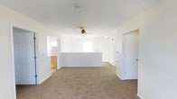 This single-story space features an open-concept layout with a bright, neutral interior defined by white walls and warm beige carpeting throughout. The main living area is generously sized, providing ample room for flexible furniture arrangements. A low half-wall or peninsula counter serves as a natural divider between the living area and an adjacent space, likely a kitchen or dining zone, visible through a pass-through opening on the left side. This architectural detail helps define separate functional zones while maintaining an open, connected feel. A ceiling fan with a light fixture is centrally mounted, contributing to both air circulation and ambient lighting. Natural light enters through a window positioned near the back wall, brightening the interior. Multiple doorways are visible, suggesting access to additional rooms beyond the main living area. The space maintains a clean, move-in ready condition with a straightforward, practical layout well-suited for everyday living. The consistent use of white throughout the walls and trim gives the interior a cohesive and airy appearance.