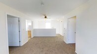 This single-story space features an open-concept layout with a bright, neutral interior defined by white walls and warm beige carpeting throughout. The main living area is generously sized, providing ample room for flexible furniture arrangements. A low half-wall or peninsula counter serves as a natural divider between the living area and an adjacent space, likely a kitchen or dining zone, visible through a pass-through opening on the left side. This architectural detail helps define separate functional zones while maintaining an open, connected feel. A ceiling fan with a light fixture is centrally mounted, contributing to both air circulation and ambient lighting. Natural light enters through a window positioned near the back wall, brightening the interior. Multiple doorways are visible, suggesting access to additional rooms beyond the main living area. The space maintains a clean, move-in ready condition with a straightforward, practical layout well-suited for everyday living. The consistent use of white throughout the walls and trim gives the interior a cohesive and airy appearance.
