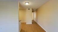 This single-floor space offers a straightforward and functional layout designed for comfortable everyday living. The interior is characterized by warm, light-toned hardwood-style flooring that runs throughout, providing a cohesive and inviting feel underfoot. Neutral, cream-colored walls complement the flooring and contribute to a bright, clean aesthetic across the space.

A central hallway serves as the main artery of the layout, connecting the various areas of the home in a linear, efficient manner. Structural columns and partial walls define the corridor while maintaining an open sightline through the length of the space. Flush-mount ceiling light fixtures provide consistent illumination throughout the hallway and adjoining areas.

The textured ceiling adds a subtle design detail, and HVAC vents are thoughtfully integrated into both the ceiling and walls to ensure climate comfort throughout. White baseboards provide a clean contrast against the warm flooring, adding a polished finish to each room transition.

Overall, the space presents a well-maintained, move-in-ready interior with a neutral palette that offers flexibility for a variety of furnishing styles. The connected layout promotes ease of movement, and the consistent use of materials throughout creates a unified, harmonious living environment.