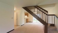 This two-floor residential space offers a practical and functional layout with a welcoming main level that sets a comfortable tone throughout. The main floor features a spacious carpeted living area that flows openly, creating a sense of generous square footage. A prominent staircase with a dark wood railing serves as a visual anchor in the space, connecting the main level to the upper floor in a straightforward and efficient design. Beneath the staircase, the layout makes clever use of the available space.

The kitchen is conveniently situated on the main level, featuring warm wood-toned cabinetry, a countertop workspace, and standard appliances including a range and overhead ventilation. The kitchen's placement adjacent to the living area promotes an easy, connected flow between the two spaces.

Natural light is a notable feature of the main floor, with a large sliding glass door that opens to an outdoor green area, bringing in brightness and providing direct access to the exterior. This element enhances the overall openness of the living space.

The neutral, light-toned walls and carpeted flooring throughout contribute to a clean and understated aesthetic. With two floors of living space, this property offers ample room for a variety of uses and configurations.
