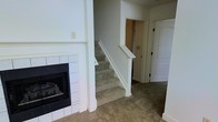 This two-story home offers a well-organized layout spread across two levels, connected by a carpeted staircase with a clean white-painted handrail and newel post. The main living area is anchored by a tile-surround fireplace with a classic white mantel, providing a focal point that adds warmth and character to the space. The flooring throughout the main level features wall-to-wall carpet in a neutral tone, contributing to a comfortable and understated aesthetic. The interior is finished in a crisp, all-white color palette across the walls, trim, and doors, creating a bright and clean atmosphere throughout. Panel-style doors and simple, functional hardware carry a traditional design sensibility consistent across the visible rooms. A hallway adjacent to the staircase provides access to additional rooms, with closet space also visible, suggesting practical storage solutions integrated into the floor plan. The overall ambiance is calm and move-in ready, with a neutral canvas that lends itself well to a variety of furnishing styles. The two-floor layout allows for a natural separation between living and private spaces, making this home both functional and versatile.