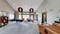 This single-floor space is designed with a mid-century modern aesthetic that balances functionality and style throughout. The main lounge area is the focal point of the space, featuring soaring vaulted ceilings that lend an open, airy feel. Two striking spherical wood pendant lights hang overhead, complementing the recessed lighting and adding a warm, sculptural element to the room. Patterned carpet in soft grey tones covers the floor, providing a neutral foundation for the bold furnishings, which include tufted blue sofas, cognac leather accent chairs, and a wood-framed glass coffee table arranged into an inviting conversation area. Large dark-trimmed windows and glass doors along one wall flood the space with natural light and offer views of an outdoor deck and surrounding greenery, further enhancing the open ambiance. A dedicated reception or workspace is positioned near the entrance, complete with a dark wood desk and leather seating. Adjacent to the main lounge, a well-appointed kitchenette features floor-to-ceiling dark cabinetry, stainless steel appliances, a gas range, dark countertops, open floating shelves, an undermount sink, and a beverage refrigerator, making the space well-suited for gatherings and everyday use.